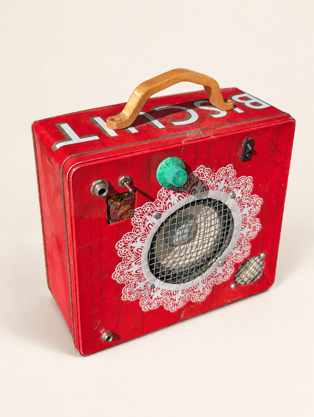 Angled view of the red biscuit tin amplifier with wooden handle, green control knob and speaker grilles against an off-white background