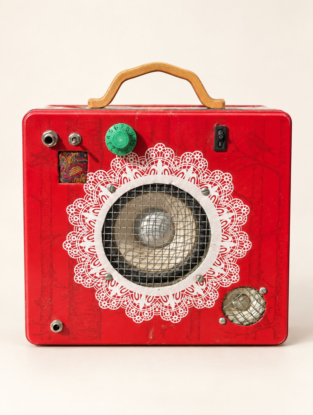 Straight-on view of the biscuit tin amplifier showing its speaker, controls and decorative red front against an off-white background